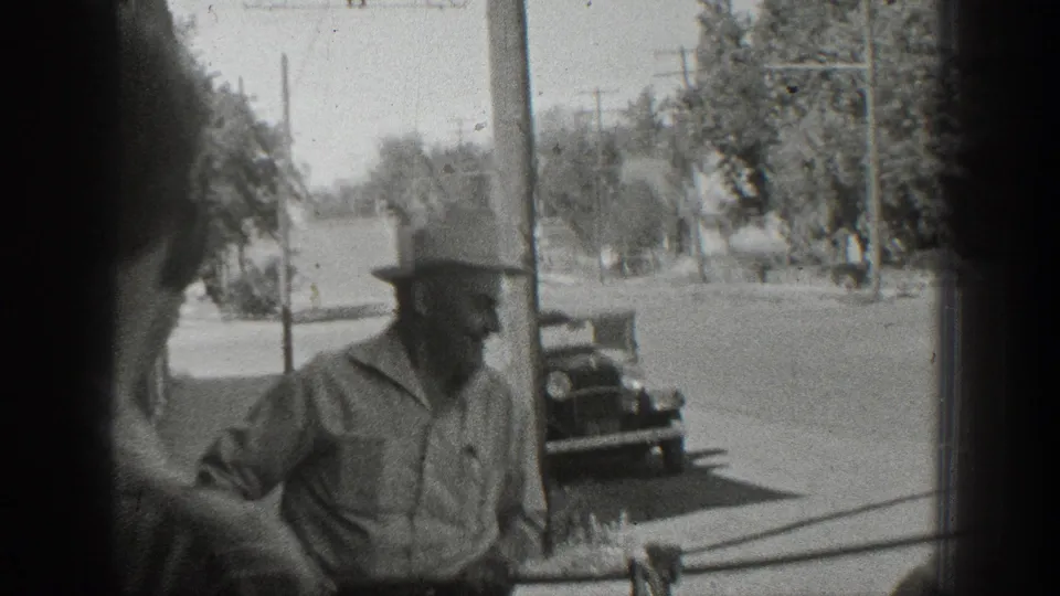 Black-and-white home-movie frame from 1936 California showing a man beside a suspended large fish on a sidewalk with cars in the background.