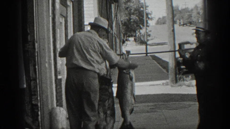 1936 California home-movie frame showing a man in a light shirt and hat handling a large fish outside a street-front building.