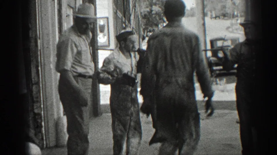 Black-and-white 1936 California frame showing several men on a sidewalk around a suspended fish, with a storefront and parked car nearby.