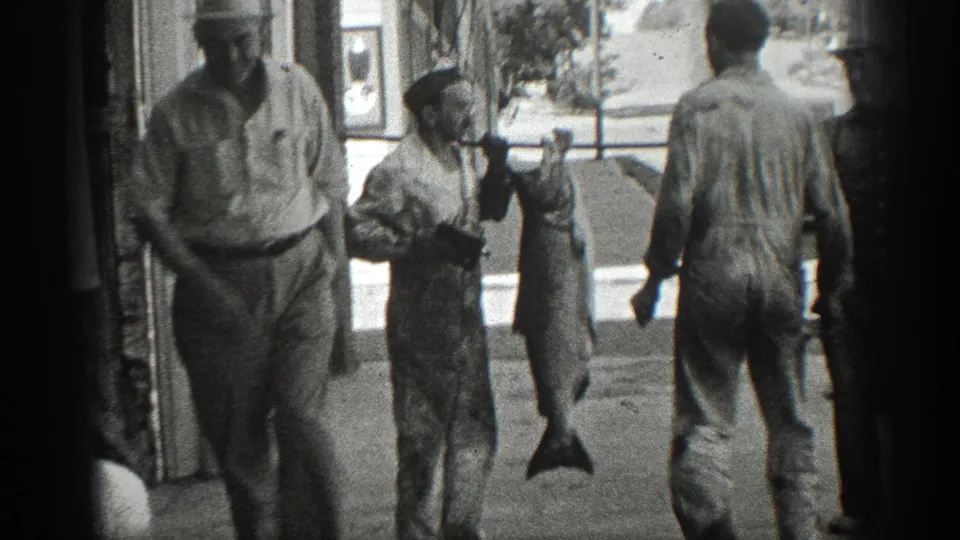 1936 California home-movie frame showing men in work clothes outside a storefront with a large fish hanging from a carrying pole.