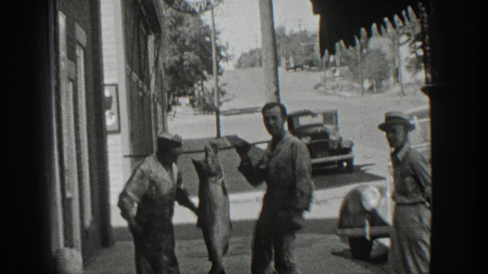 Black-and-white 8mm frame from 1936 California showing men on a sidewalk with a large fish suspended between them.
