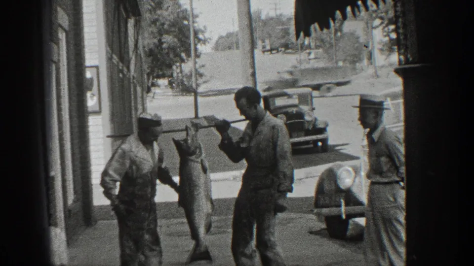 1936 California home-movie frame showing several men around a large fish displayed on a pole near a storefront and vintage cars.