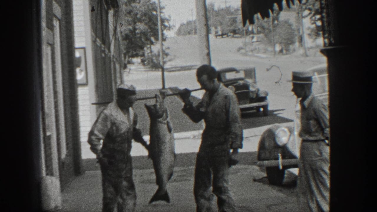 Men on a sidewalk hold a large fish on a pole near storefronts and parked cars.