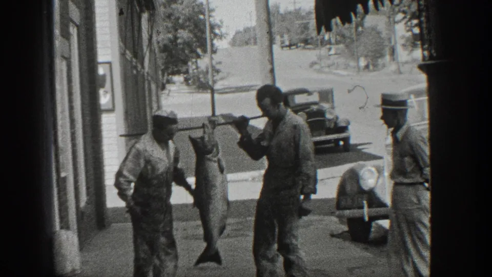 Black-and-white 1936 California home-movie frame showing men on a town sidewalk displaying a large fish suspended from a pole, with storefronts and early automobiles nearby.
