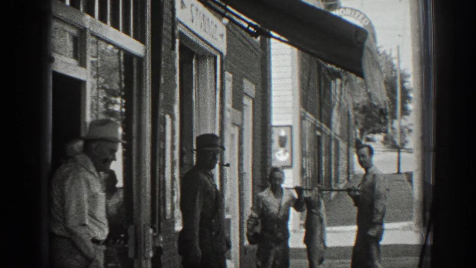 Black-and-white 1936 California street frame with men outside storefronts, one large fish hanging from a pole, and bystanders positioned along the sidewalk.