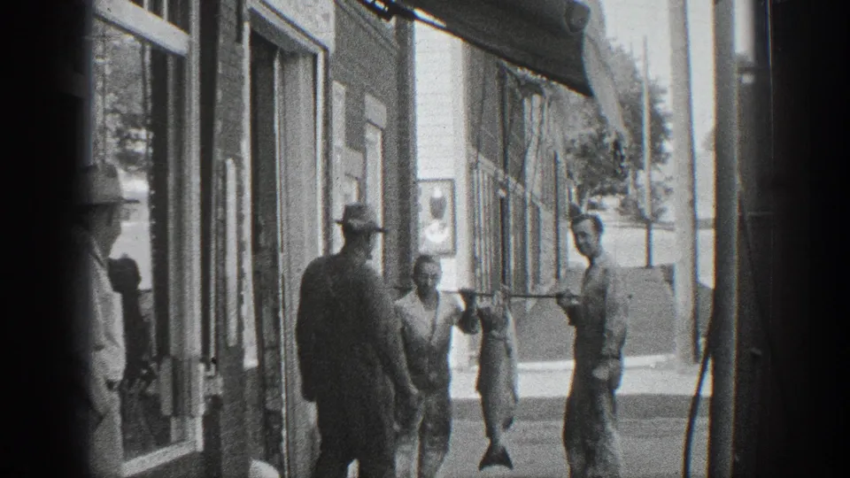 Black-and-white 1936 California home-movie frame showing the sidewalk group from farther back, with storefront windows, awnings, and the large fish carried between men.