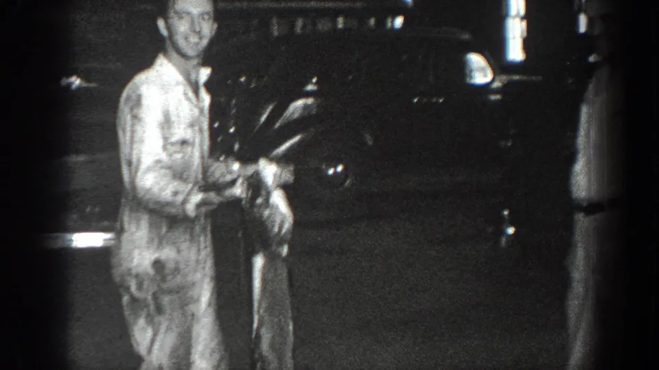 Black-and-white 1936 California home-movie frame showing a smiling man near a dark parked automobile with the large fish visible beside him.