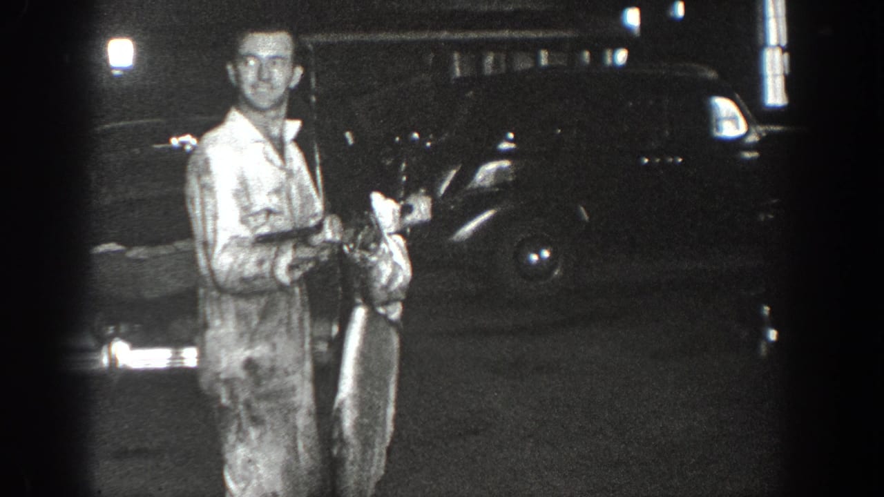 Man standing in a garage area holding an unclear object near a dark car.