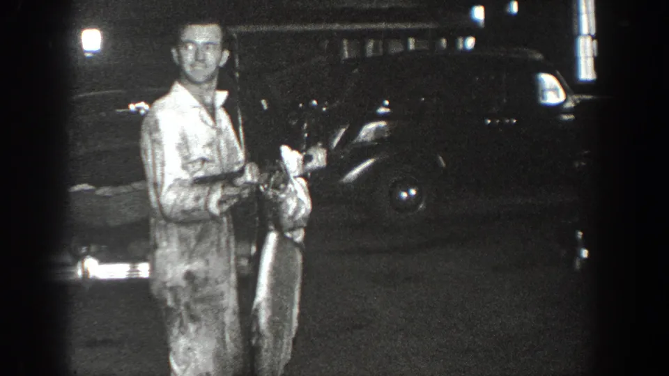 In 1936 California home-movie footage, a man stands in a garage-like space holding an unclear object beside a dark period car.