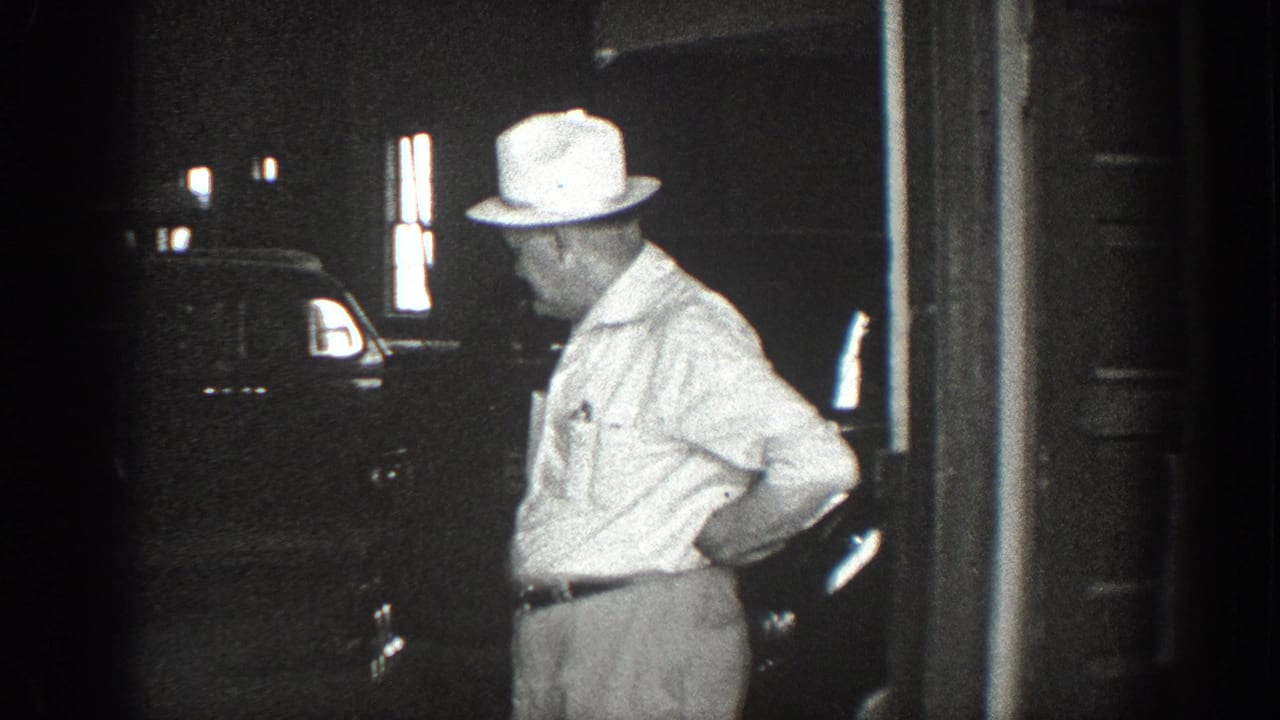 A man in a light brimmed hat stands near a dark car in a garage or doorway area in 1936 California footage.
