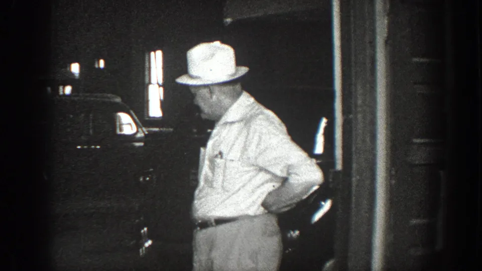 A man in a light brimmed hat stands near a dark car in a garage or doorway area in 1936 California footage.