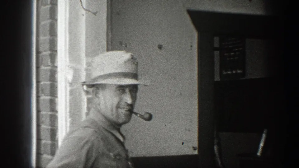 A man in a cap smokes a pipe beside a building entrance in the same 1936 California home-movie sequence.