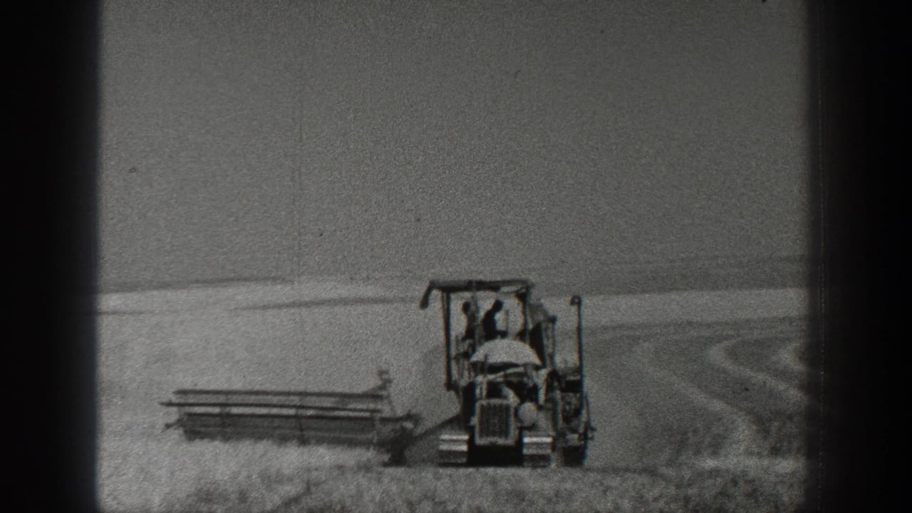 Black-and-white 8mm view of a farmer on a tractor pulling harvesting equipment in rural California.