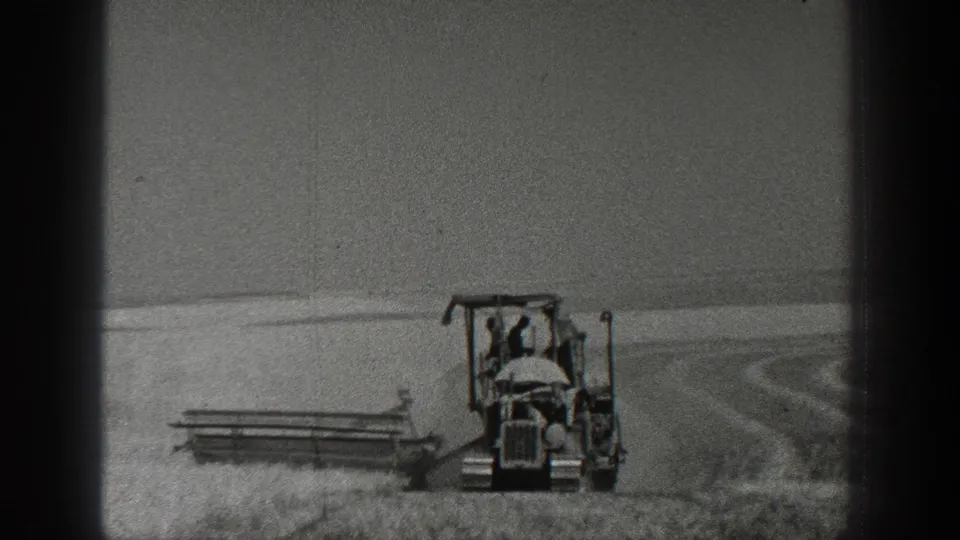 Black-and-white 8mm view of a farmer on a tractor pulling harvesting equipment in rural California.