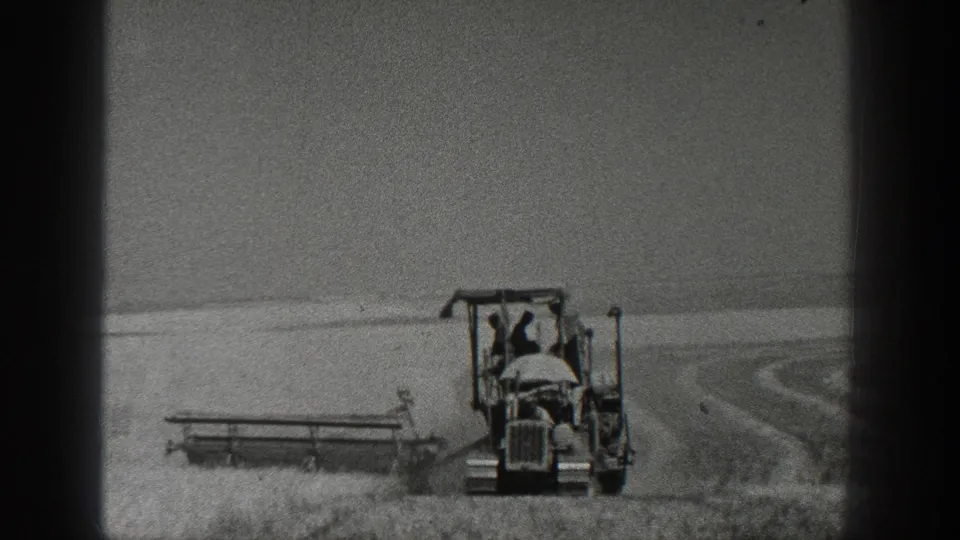 1930s home-movie frame of a tractor harvester moving through a grain field, with the operator visible at distance.