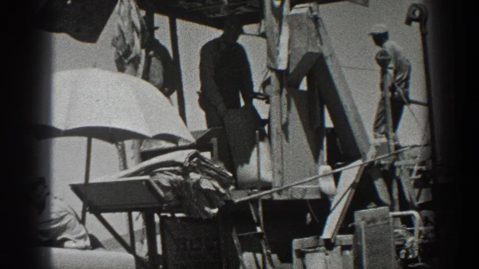 Men in work clothes operate or tend a 1936 California combine harvester, with a large umbrella shading part of the machinery.