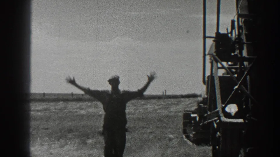 In 1936 California farm footage, a man in a cap stands near large field equipment and raises both arms against a flat horizon.