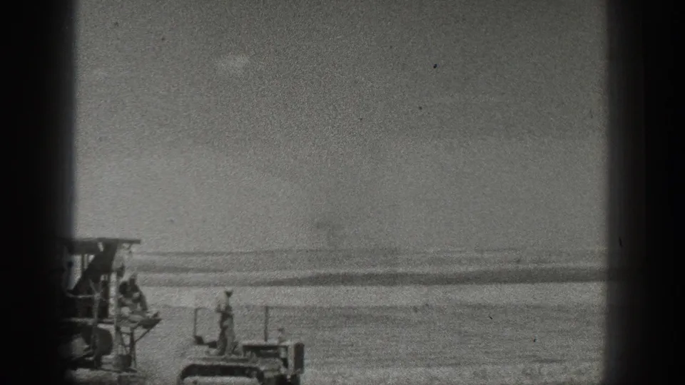 A distant worker and farm equipment appear small against a broad California field and flat horizon in black-and-white 8mm footage.