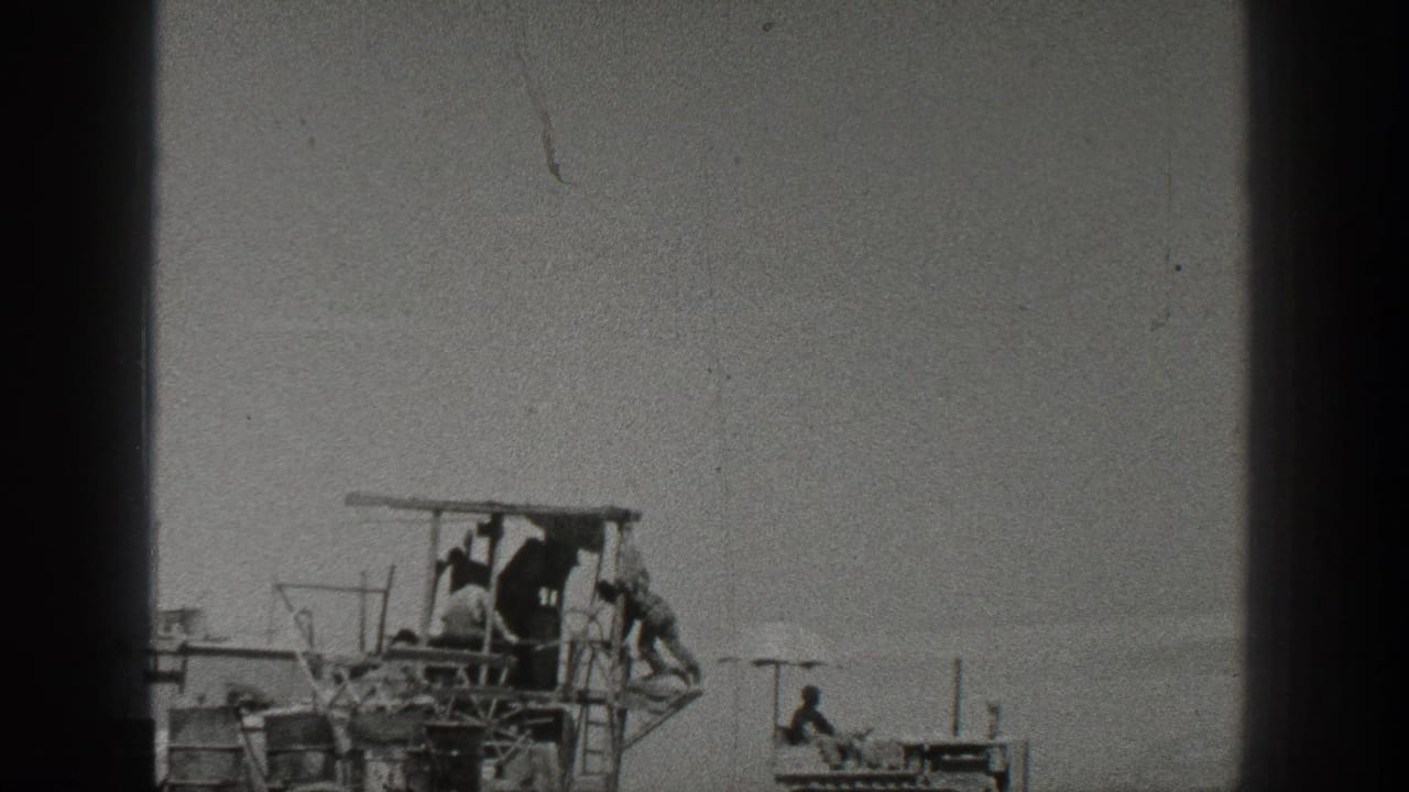 1936 California home-movie frame showing workers on a harvesting machine with a small crawler tractor nearby in a dusty open field.