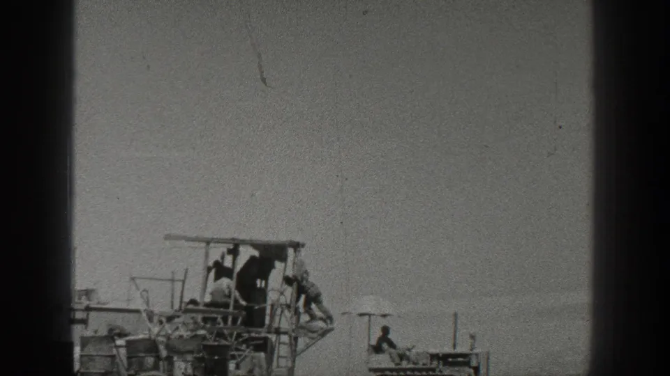 1936 California home-movie frame showing workers on a harvesting machine with a small crawler tractor nearby in a dusty open field.