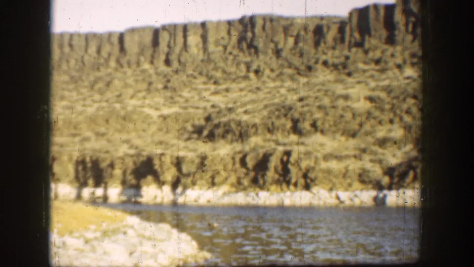 1936 California 8mm frame showing a river bend, pale rocky shore, and layered dry badland slopes in warm light.