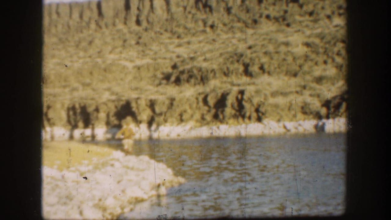 1936 California home-movie frame showing a small adult figure near a rocky shoreline, calm water, cliff face, and dark window-frame edges.