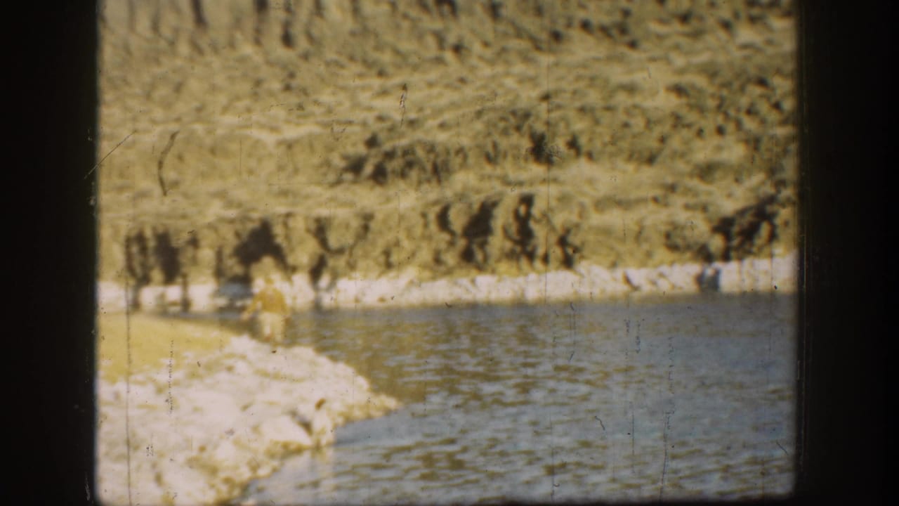 California coastal frame from 1936 showing a distant adult on a shoreline beside calm water and a steep, textured cliff.