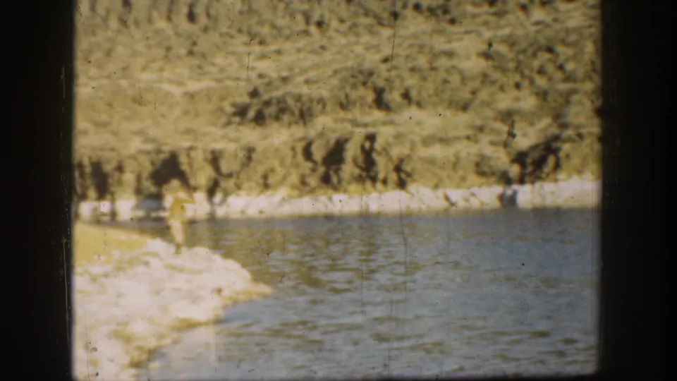 1936 California home-movie still with a lone adult walking along a rocky beach, with calm water and cliff textures filling the frame.