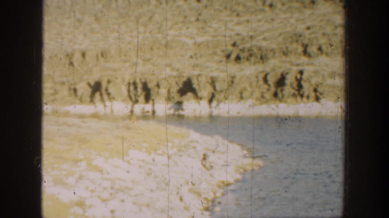 1936 California 8mm frame of a curved rocky shoreline beside blue water and a sunlit cliff or hillside in the background.