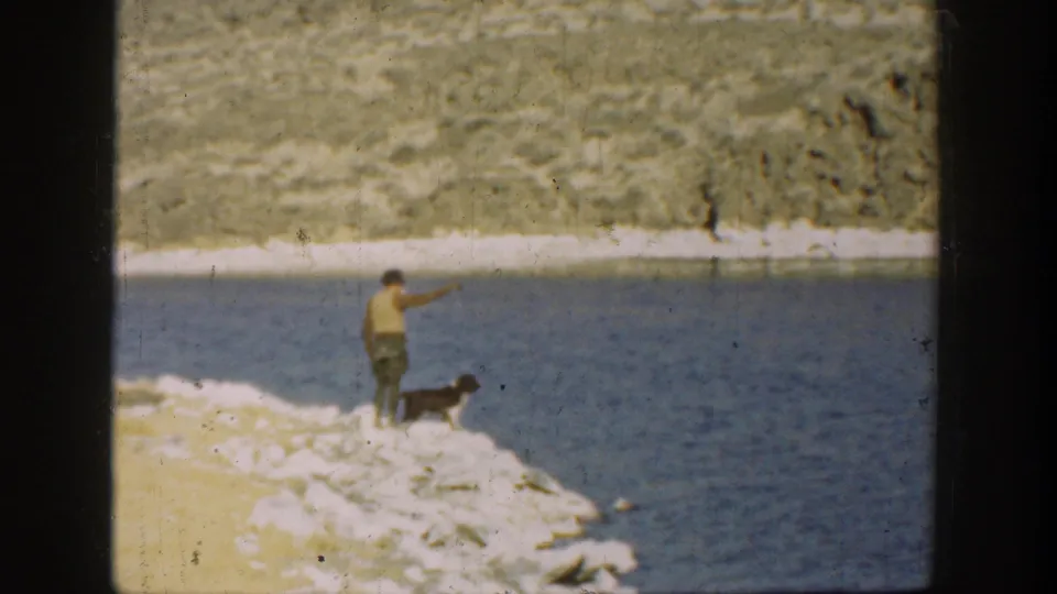 1936 California home-movie frame showing a man with a dark dog on a rocky shoreline, looking or pointing across the water.