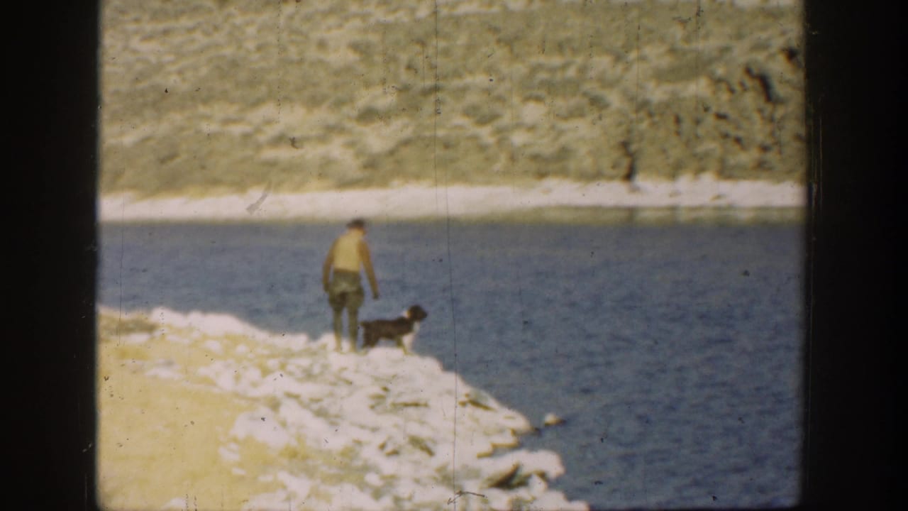 1936 California 8mm frame showing a young boy and black-and-white dog paused on a rocky lakeshore with dry hills beyond the lake.