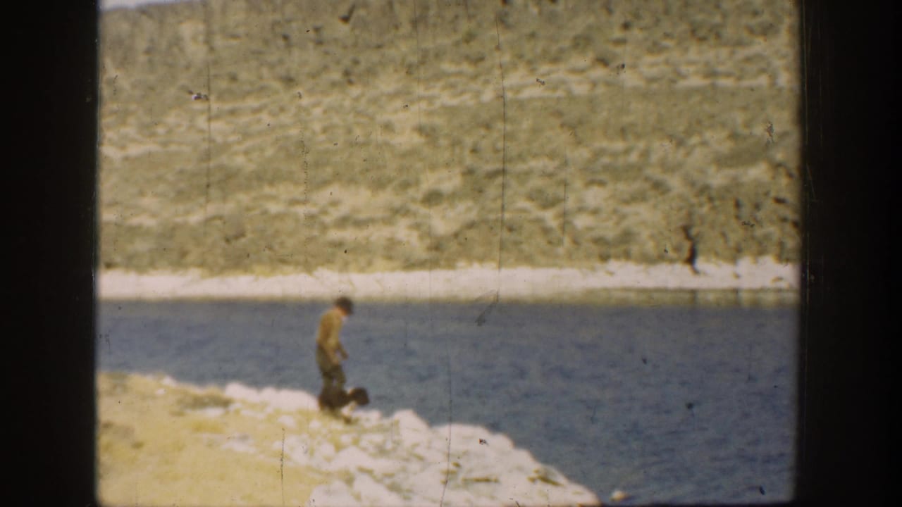 1936 California home-movie frame with a boy and dog moving along a pale rocky shoreline beside calm blue lake water.