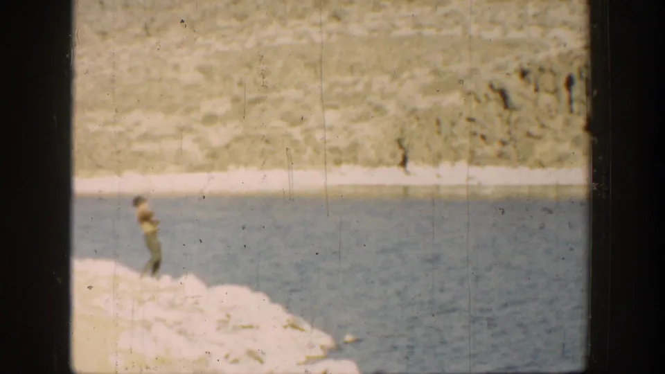1936 California 8mm frame showing the boy on a sunlit rocky lakeshore, with the dog out of view and blue lake water behind him.