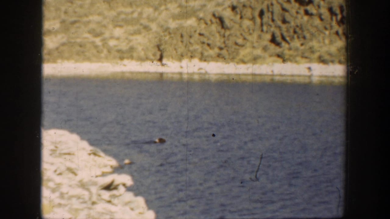 1936 California 8mm frame of a pale rocky river edge, blue water, and scrubby dry terrain in the distance.