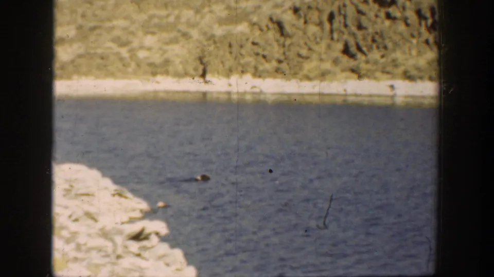 1936 California 8mm frame of a pale rocky river edge, blue water, and scrubby dry terrain in the distance.