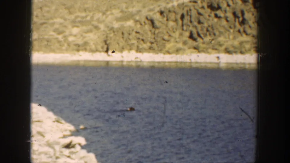 1936 California 8mm frame showing rippled blue river water, pale rocks, and a dry hillside across the channel.