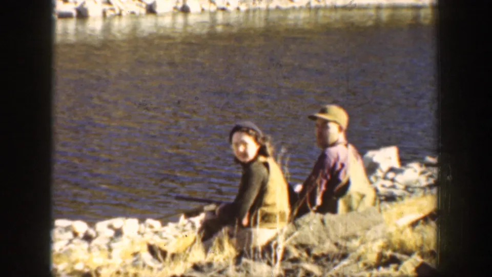 A couple in 1930s attire sits on a rocky California riverside bank, with water and hillside behind them.