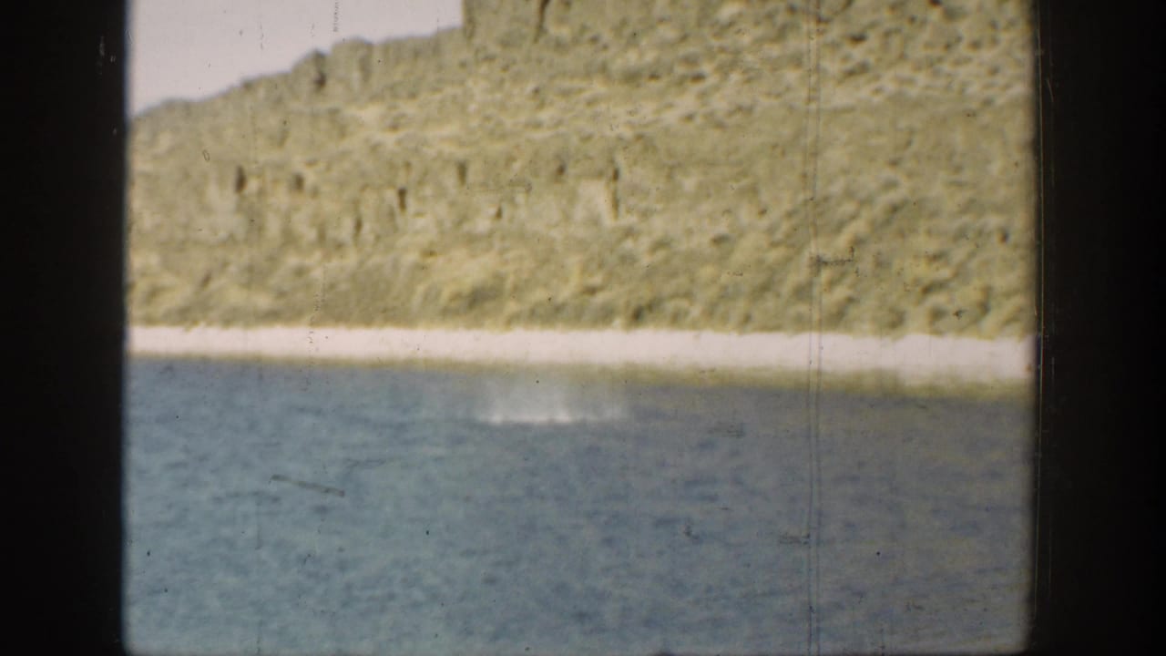 1936 California 8mm frame with blue water in the foreground and a rocky sunlit bank across the shore.