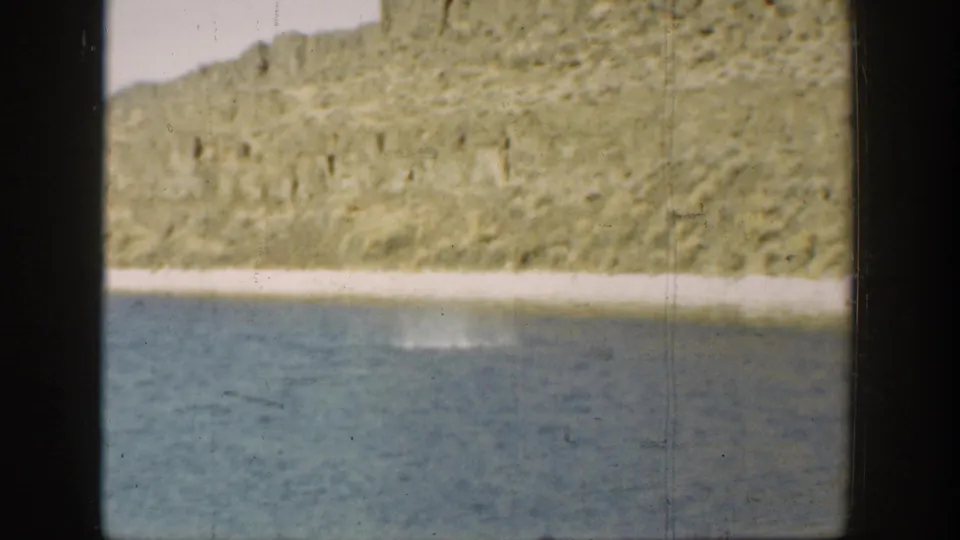 1936 California 8mm frame with blue water in the foreground and a rocky sunlit bank across the shore.
