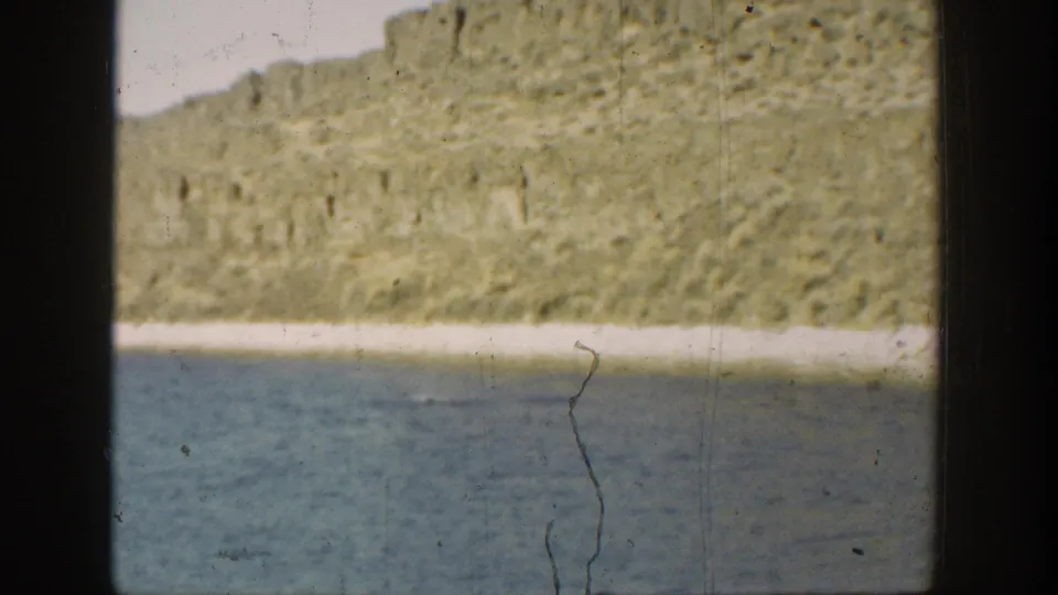 1936 California home-movie still showing water, a pale shore strip, and a rocky hillside across the river or lake.