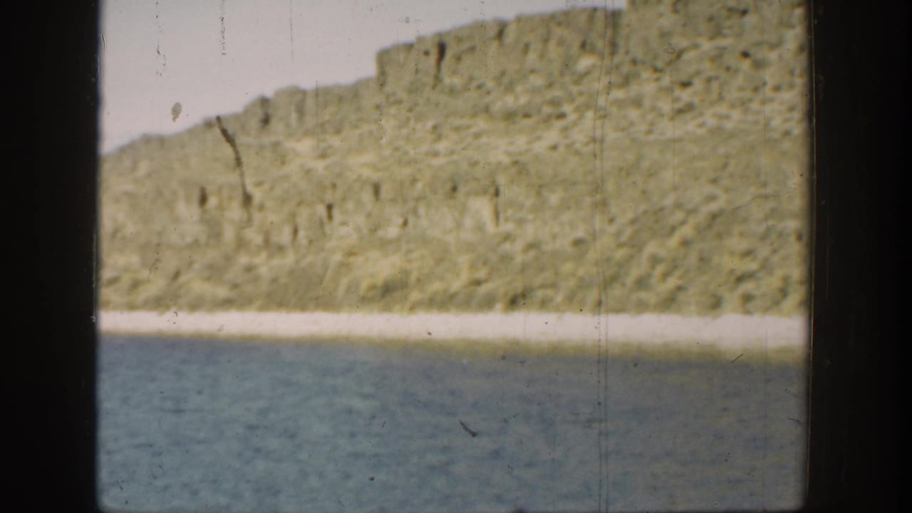 1936 California 8mm view across lagoon water toward a rugged, sunlit cliff and narrow pale shore.