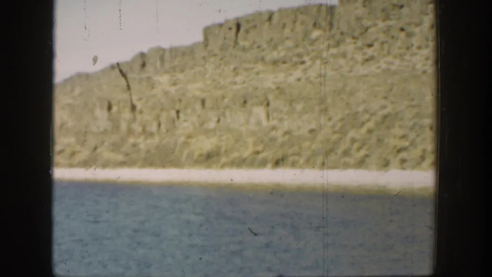 1936 California 8mm view across lagoon water toward a rugged, sunlit cliff and narrow pale shore.