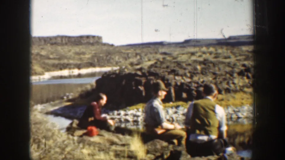 1936 California home-movie frame of three men seated by a rocky lagoon shore with water, cliffs, and scrub vegetation behind them.