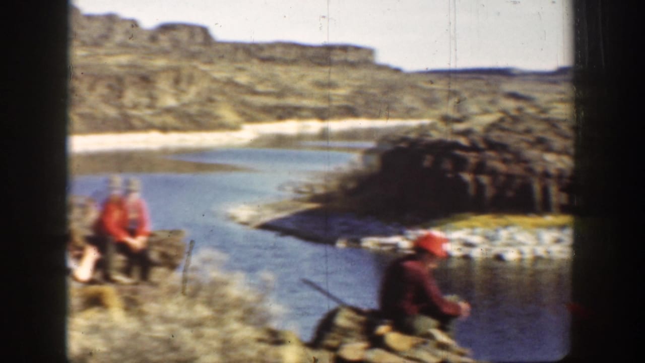 1936 California home-movie frame showing two men in red hats on a rocky riverbank, with one man holding a long gun.