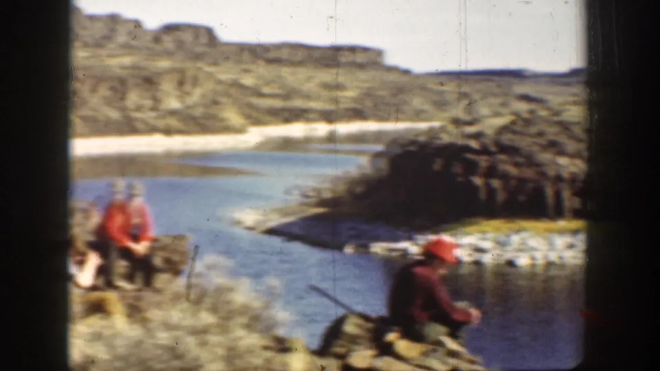 1936 California home-movie frame showing two men in red hats on a rocky riverbank, with one man holding a long gun.