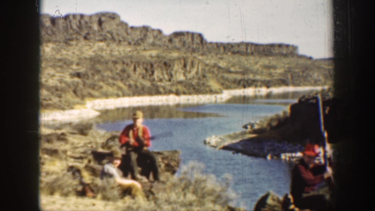 Men sit on a rocky riverbank while another person with a gun appears at the right edge.