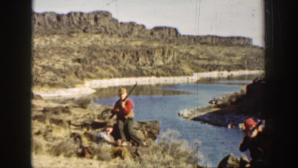 1936 California frame showing a hunter standing with a long gun while others sit beside a calm river in arid terrain.