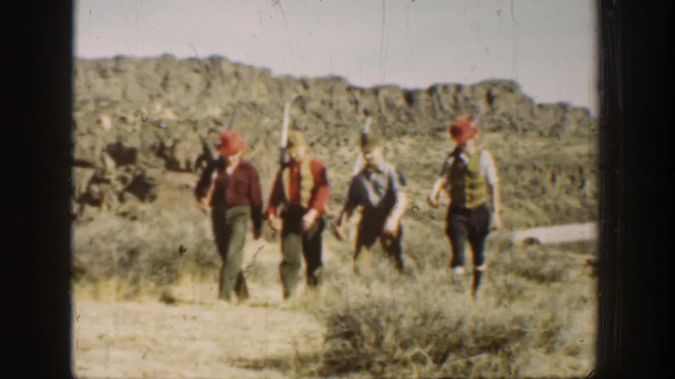 Four men in hats and casual period clothing approach the camera with long rifles in a rugged California outdoor setting.
