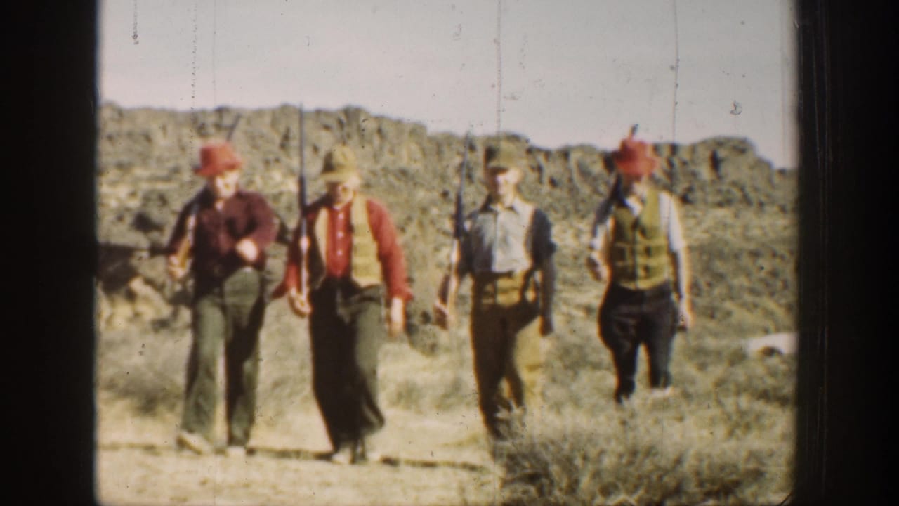 Four men in hats continue marching through a rocky desert area with rifles.