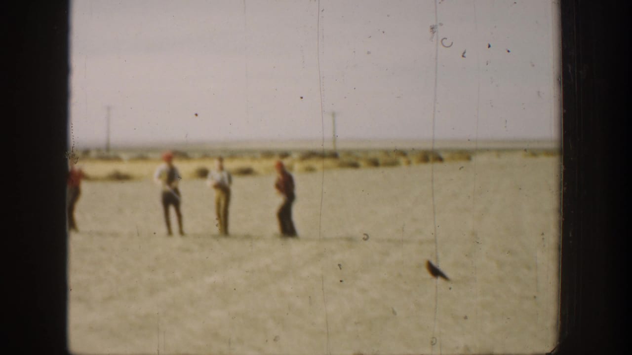 Several distant men stand in an open field with low brush and utility poles behind them.